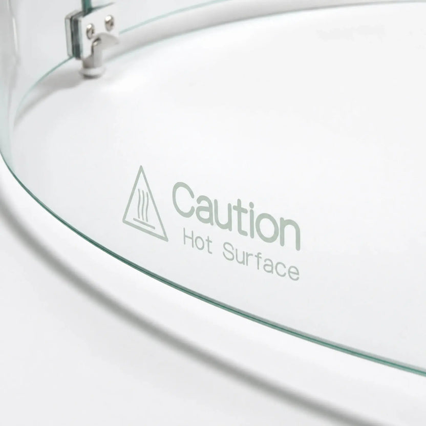 Close-up of the “Caution Hot Surface” warning printed on the Flame Havens Refuge Series round tempered glass wind guard shield. The 28-inch-diameter by 7-inch-tall accessory shows its curved clear glass edge, heat warning symbol, and part of the silver connecting hardware.