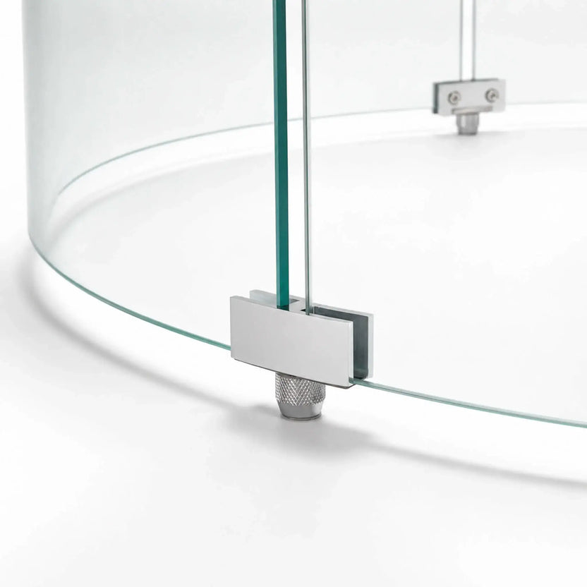 Close-up of a support foot on the Flame Havens Refuge Series 28-inch round by 7-inch-tall tempered glass wind guard shield. The silver metal bracket secures two curved glass panels while a knurled adjustable foot supports the assembly above the fire pit surface.