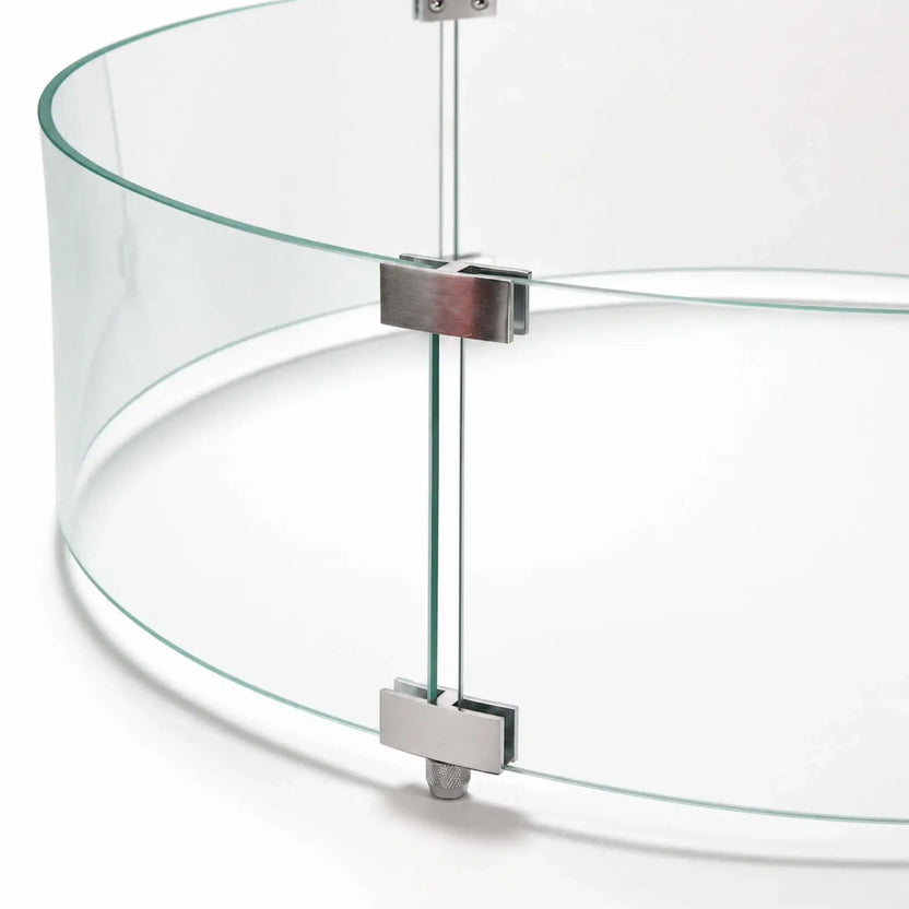 Close-up of the hardware joining the curved panels of the Flame Havens Refuge Series 28-inch round by 7-inch-tall tempered glass wind guard shield. Silver upper and lower clamps align the clear glass edges, with screws and an adjustable support foot visible against a white background.