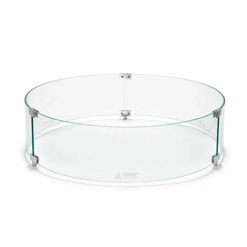Front main view of the Flame Havens Refuge Series 28-inch round by 7-inch-tall tempered glass wind guard shield. The clear circular enclosure features green-tinted glass edges, silver panel connectors, three raised support feet, and visible hot-surface caution markings along the lower panels.