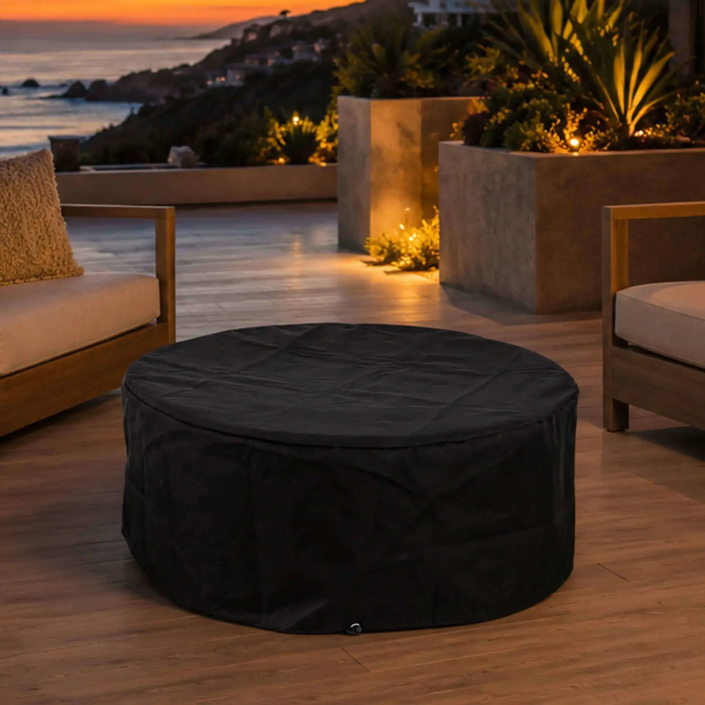 The Flame Havens Refuge Series 36-inch Round Natural Gas GFRC Concrete Fire Pit in Black displayed on a luxury outdoor patio with the custom black canvas fabric cover installed. Positioned between modern lounge seating overlooking the ocean at sunset, the image illustrates how the protective cover preserves the fire pit while complementing upscale outdoor living spaces.