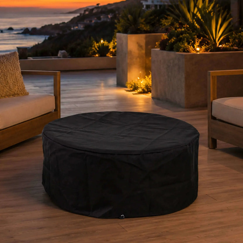The Flame Havens Refuge Series 36-inch Round Natural Gas GFRC Concrete Fire Pit in Black displayed on a luxury outdoor patio with the custom black canvas fabric cover installed. Positioned between modern lounge seating overlooking the ocean at sunset, the image illustrates how the protective cover preserves the fire pit while complementing upscale outdoor living spaces.