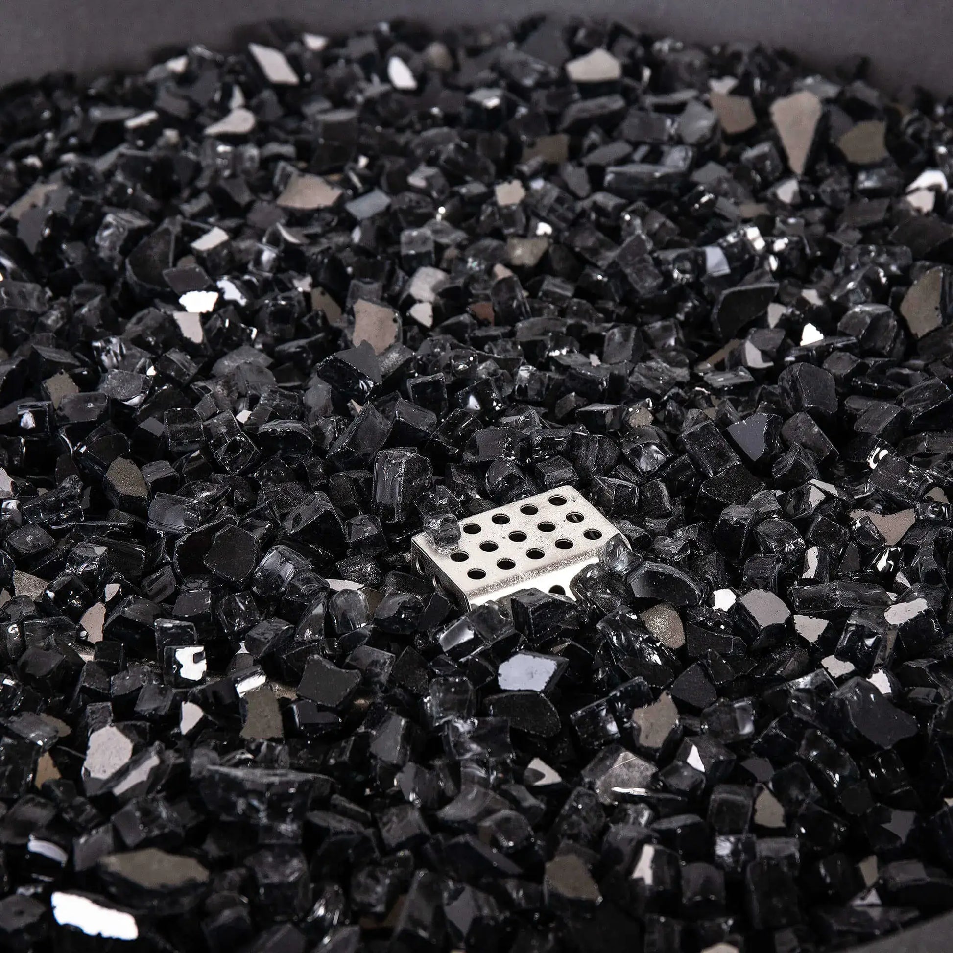 Detailed close-up of the Flame Havens Refuge Series 36-inch Round Natural Gas GFRC Concrete Fire Pit featuring premium black reflective fire glass surrounding the stainless steel CSA ignition assembly. The image emphasizes the glass texture, reflective finish, and clean installation that enhances both daytime appearance and nighttime flame presentation.