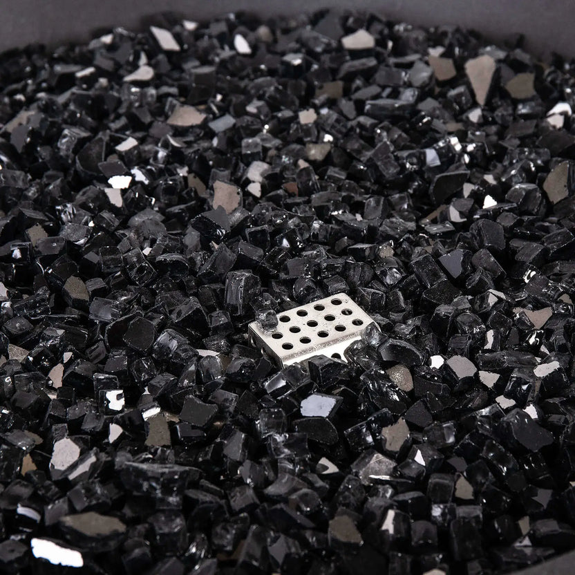 Detailed close-up of the Flame Havens Refuge Series 36-inch Round Natural Gas GFRC Concrete Fire Pit featuring premium black reflective fire glass surrounding the stainless steel CSA ignition assembly. The image emphasizes the glass texture, reflective finish, and clean installation that enhances both daytime appearance and nighttime flame presentation.