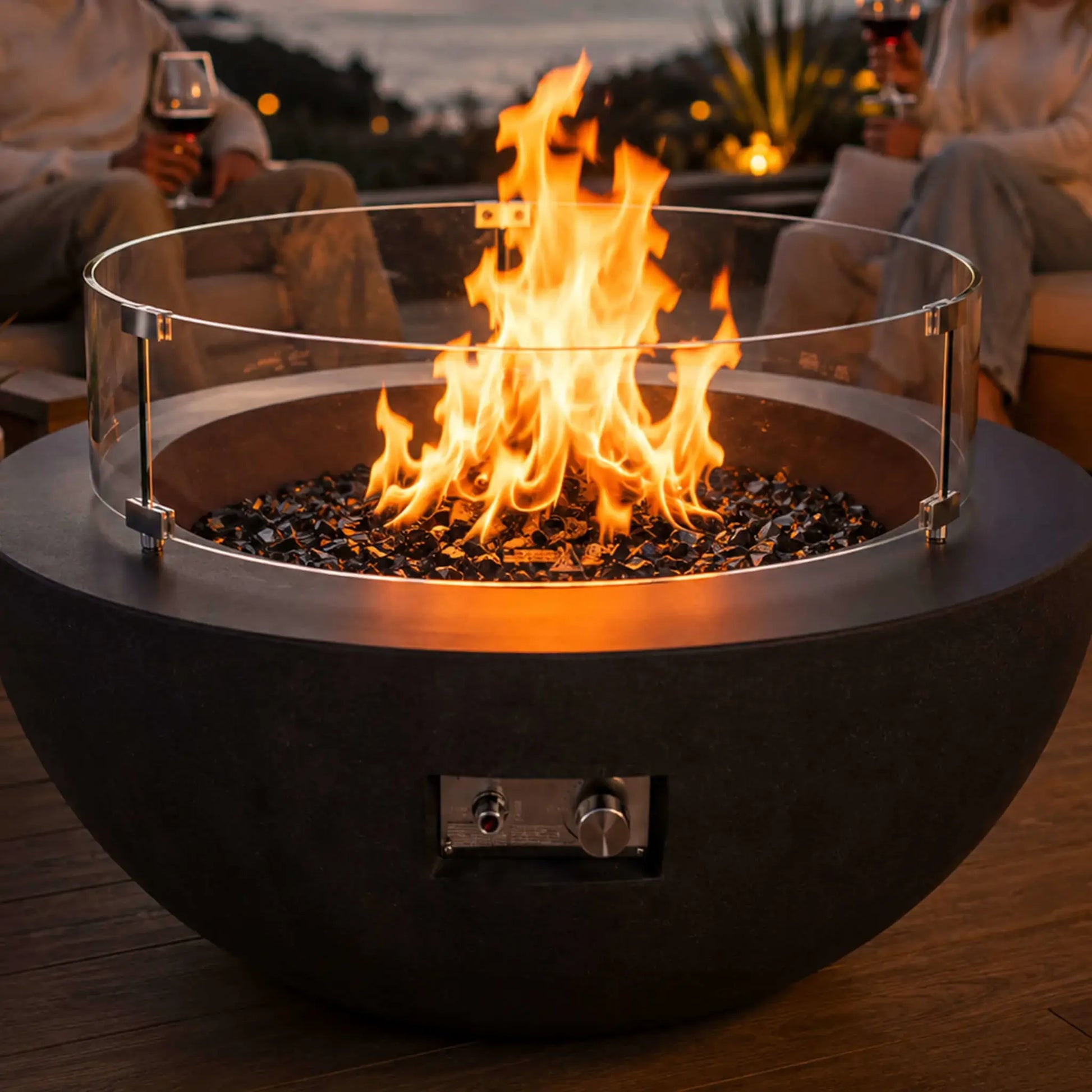 Lifestyle image of the Flame Havens Refuge Series 36-inch Round Natural Gas GFRC Concrete Fire Pit in Black featuring premium tempered black reflective fire glass and the optional tempered glass wind guard. Positioned on a luxury oceanfront patio, the fire pit creates a warm focal point as guests relax with drinks during sunset, illustrating upscale outdoor entertaining.