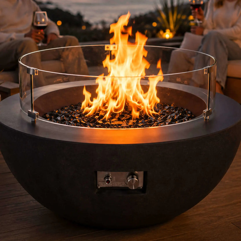 Lifestyle image of the Flame Havens Refuge Series 36-inch Round Natural Gas GFRC Concrete Fire Pit in Black featuring premium tempered black reflective fire glass and the optional tempered glass wind guard. Positioned on a luxury oceanfront patio, the fire pit creates a warm focal point as guests relax with drinks during sunset, illustrating upscale outdoor entertaining.
