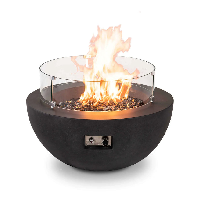 Lifestyle image of the Flame Havens Refuge Series 36-inch Round Natural Gas GFRC Concrete Fire Pit in Black featuring premium tempered black reflective fire glass and the optional tempered glass wind guard. Positioned on a luxury oceanfront patio, the fire pit creates a warm focal point as guests relax with drinks during sunset, illustrating upscale outdoor entertaining.
