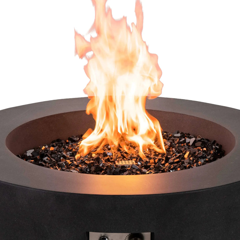 Close-up of the Flame Havens Refuge Series 36-inch Round Natural Gas GFRC Concrete Fire Pit in Black with bright natural gas flames dancing above premium tempered black reflective fire glass. The image captures the rich texture of the GFRC concrete, reflective fire media, and powerful flame display while the front ignition controls remain partially visible below.