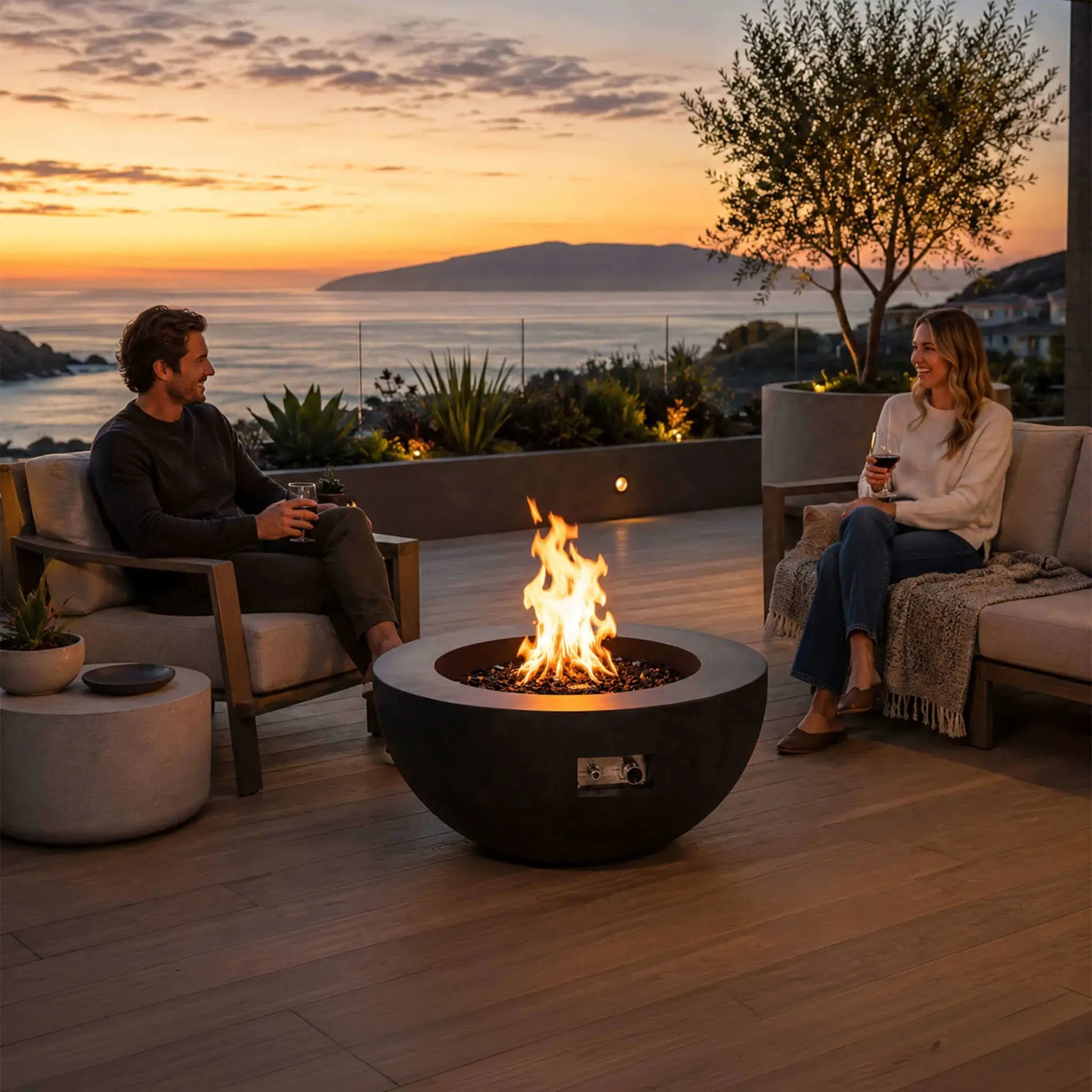 Lifestyle image of the Flame Havens Refuge Series 36-inch Round Natural Gas GFRC Concrete Fire Pit in Black burning with premium tempered black reflective fire glass on a luxury outdoor patio overlooking the ocean at sunset. Two people relax with drinks around the fire, demonstrating how the modern GFRC concrete fire pit creates a warm and inviting gathering space for outdoor entertaining.