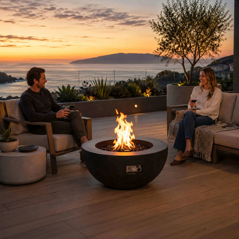 Lifestyle image of the Flame Havens Refuge Series 36-inch Round Natural Gas GFRC Concrete Fire Pit in Black burning with premium tempered black reflective fire glass on a luxury outdoor patio overlooking the ocean at sunset. Two people relax with drinks around the fire, demonstrating how the modern GFRC concrete fire pit creates a warm and inviting gathering space for outdoor entertaining.