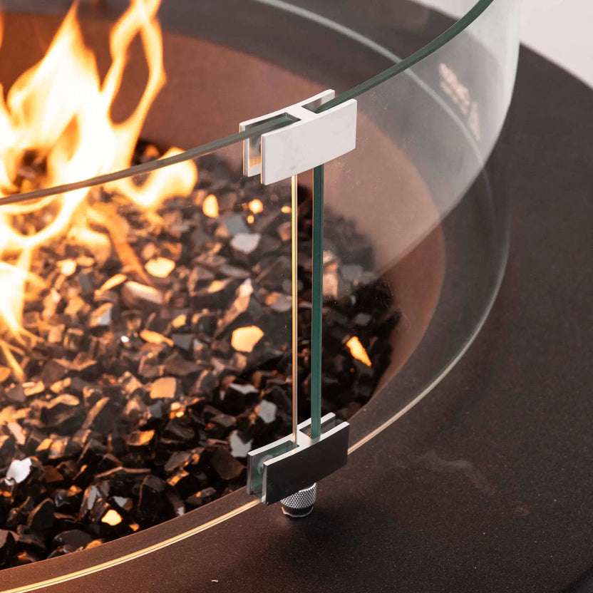 Close-up detail of the optional tempered glass wind guard installed on the Flame Havens Refuge Series 36-inch Round Natural Gas GFRC Concrete Fire Pit in Black. The image highlights the precision stainless steel mounting brackets, thick tempered glass panels, premium black reflective fire glass, and active flames, showcasing the high-quality craftsmanship and secure wind guard installation.
