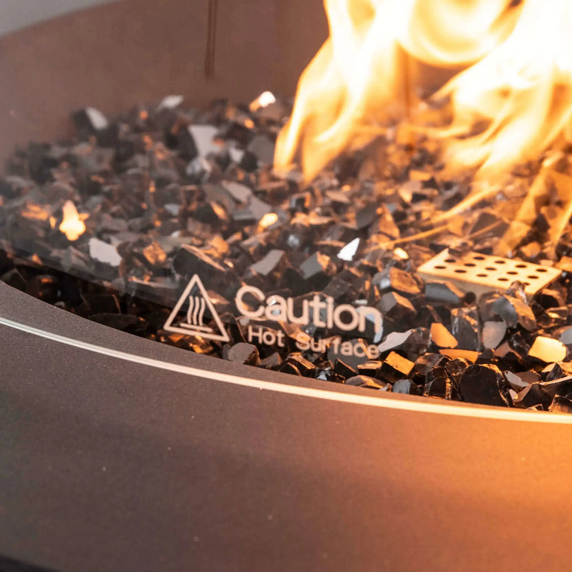 Detailed close-up of the Flame Havens Refuge Series 36-inch Round Natural Gas GFRC Concrete Fire Pit in Black featuring premium tempered black reflective fire glass, active natural gas flames, and the optional tempered glass wind guard. The etched "Caution Hot Surface" marking on the glass is clearly visible, emphasizing the safety features while highlighting the elegant flame presentation and premium outdoor construction.