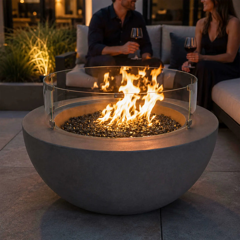 The Flame Havens Refuge Series 36-inch Round Natural Gas GFRC Concrete Fire Pit in Gray creates a welcoming focal point in a luxury outdoor patio. A clear tempered glass wind guard surrounds the lively flame over black tempered reflective fire glass while guests relax nearby on modern outdoor seating, showcasing an upscale evening entertaining environment.