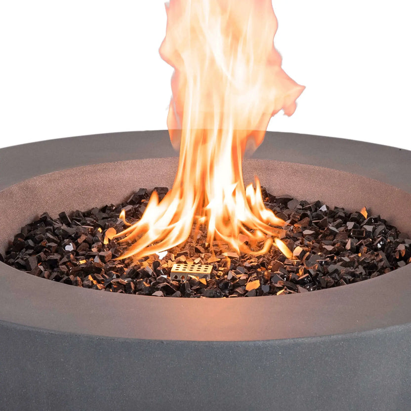 Detailed close-up of the Flame Havens Refuge Series 36-inch Round Natural Gas GFRC Concrete Fire Pit in Gray with black tempered reflective fire glass surrounding the stainless steel burner. Bright flames rise from the burner while the textured gray GFRC concrete rim frames the fire, highlighting the premium materials and realistic flame presentation.