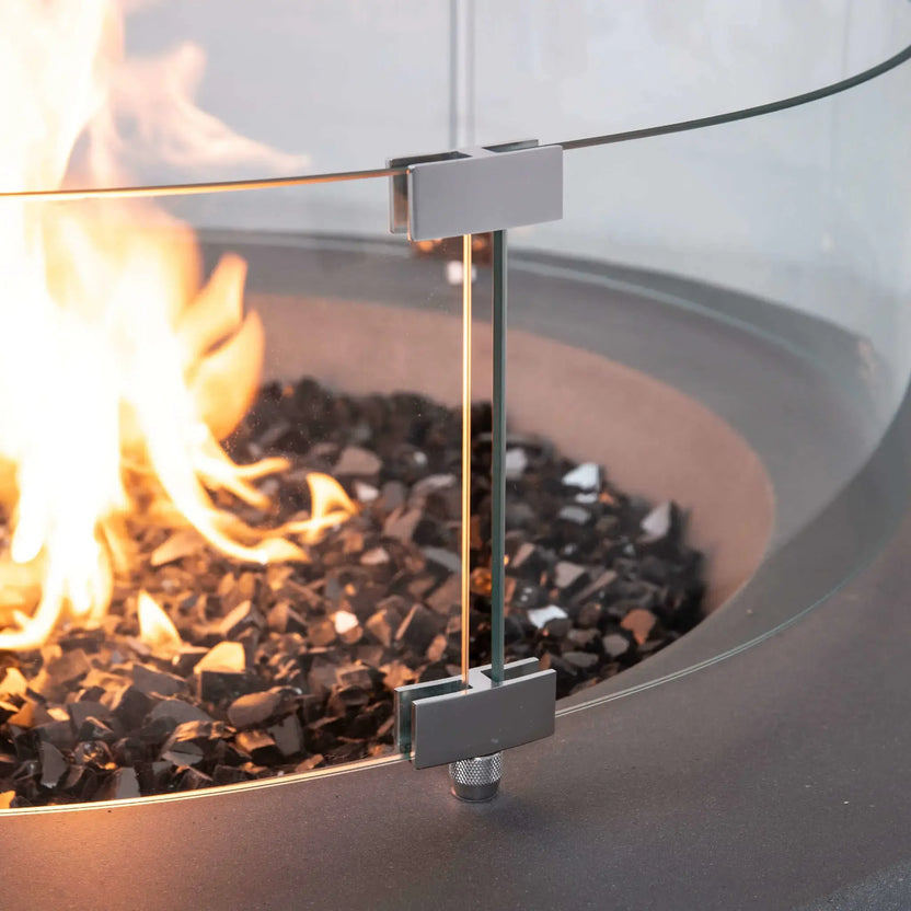 Close-up of the Flame Havens Refuge Series tempered glass wind guard mounted on the 36-inch Round Natural Gas GFRC Concrete Fire Pit in Gray. The image highlights the precision stainless steel glass mounting brackets, crystal-clear tempered safety glass, black tempered reflective fire glass, and vibrant flames, showcasing the premium craftsmanship and durable outdoor construction.