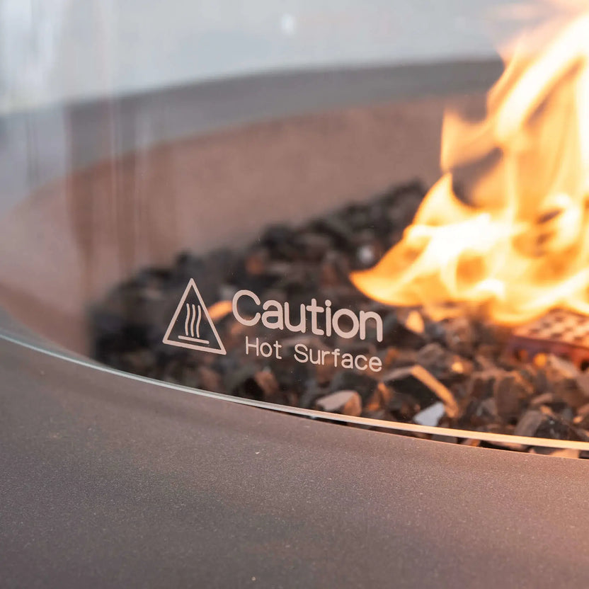 Detailed close-up of the Flame Havens Refuge Series tempered glass wind guard featuring the etched "Caution Hot Surface" safety marking. The image captures the clear tempered glass surrounding lively flames over black tempered reflective fire glass inside the 36-inch Round Natural Gas GFRC Concrete Fire Pit in Gray, emphasizing both safety and premium outdoor fire pit design.