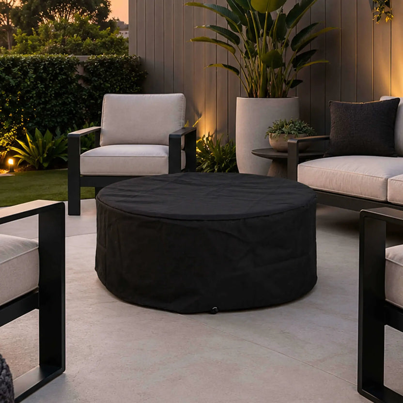 The Flame Havens Refuge Series 36-inch Round Natural Gas GFRC Concrete Fire Pit sits beneath its black canvas fabric cover in a sophisticated outdoor patio setting. Surrounded by modern lounge seating, landscaping, and ambient lighting, the image demonstrates how the weather-resistant cover protects the fire pit while complementing luxury outdoor living spaces.