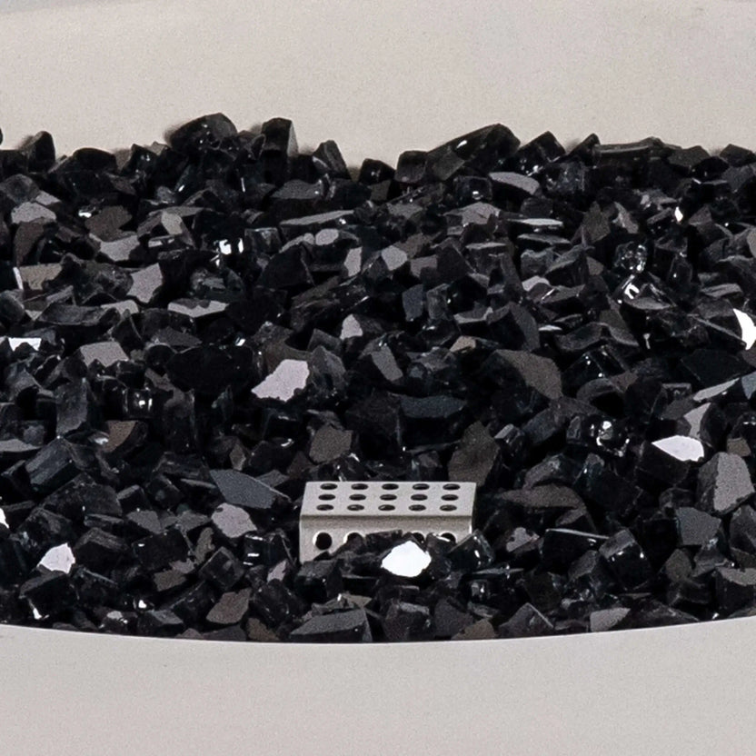 Detailed close-up of the Flame Havens Refuge Series black reflective tempered fire glass inside the 36-inch Round Natural Gas White GFRC Concrete Fire Pit. The image showcases the premium fire media surrounding the stainless steel CSA ignition system, highlighting the reflective finish and elegant appearance before the fire feature is lit.
