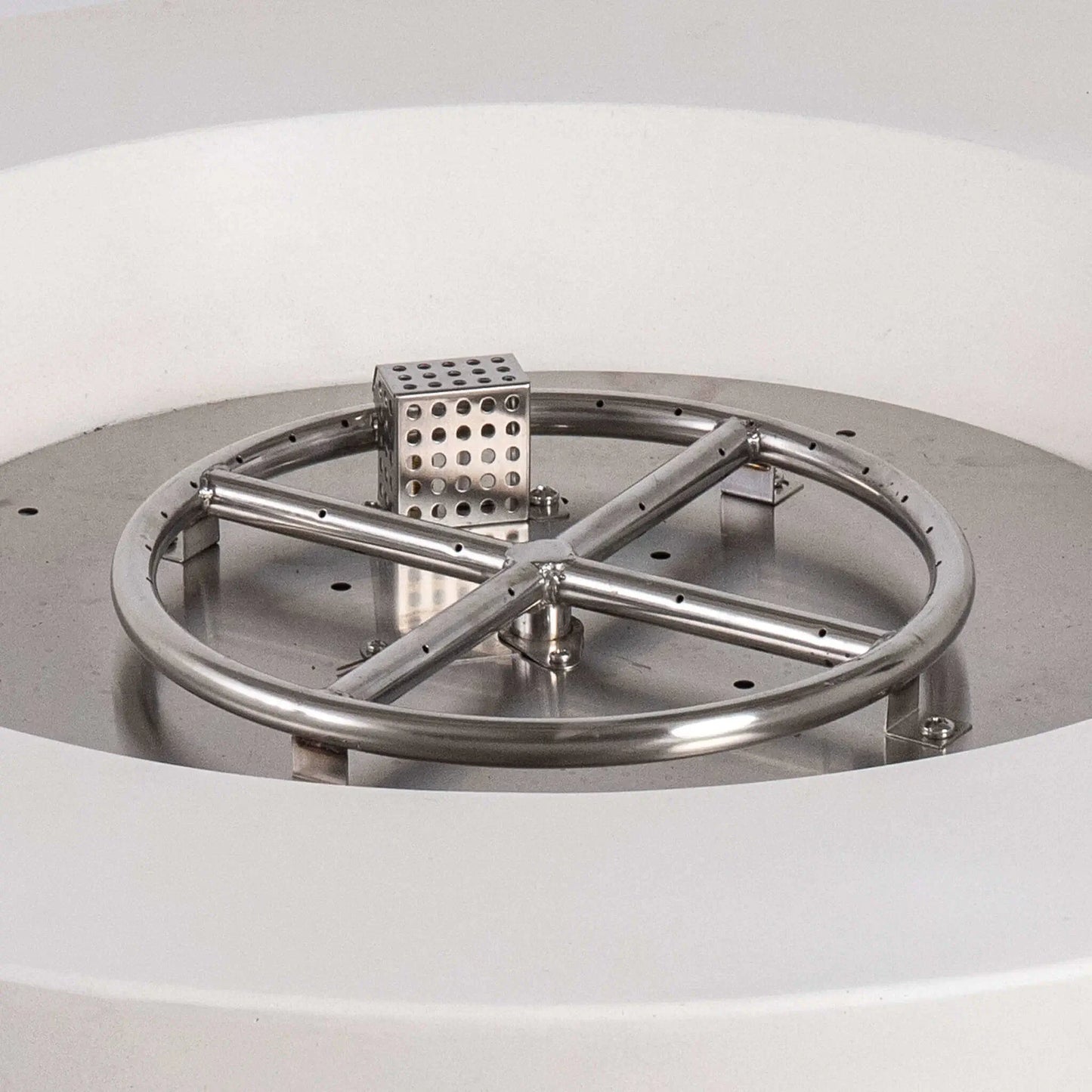 Close-up of the Flame Havens Refuge Series stainless steel burner and CSA-certified ignition assembly inside the 36-inch Round Natural Gas White GFRC Concrete Fire Pit. The precision-engineered circular burner, cross supports, and ignition housing demonstrate the premium construction designed for reliable outdoor fire performance.