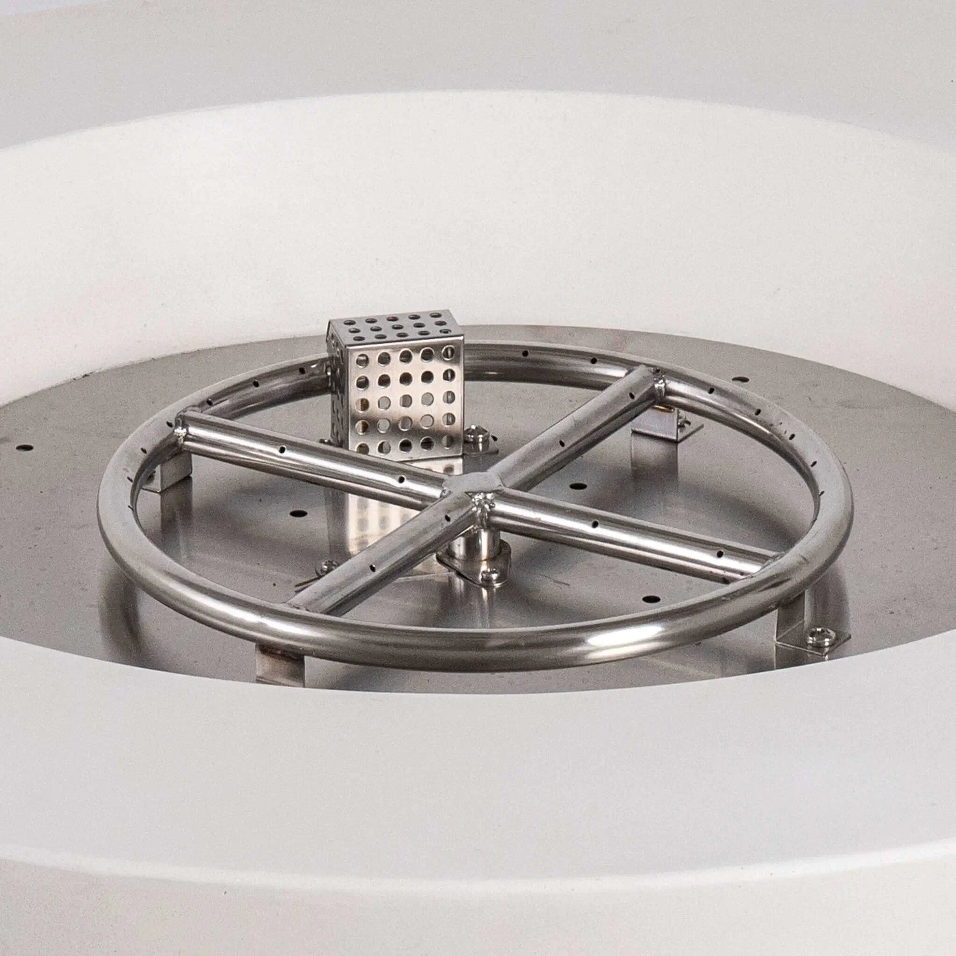 Close-up of the Flame Havens Refuge Series stainless steel burner and CSA-certified ignition assembly inside the 36-inch Round Natural Gas White GFRC Concrete Fire Pit. The precision-engineered circular burner, cross supports, and ignition housing demonstrate the premium construction designed for reliable outdoor fire performance.