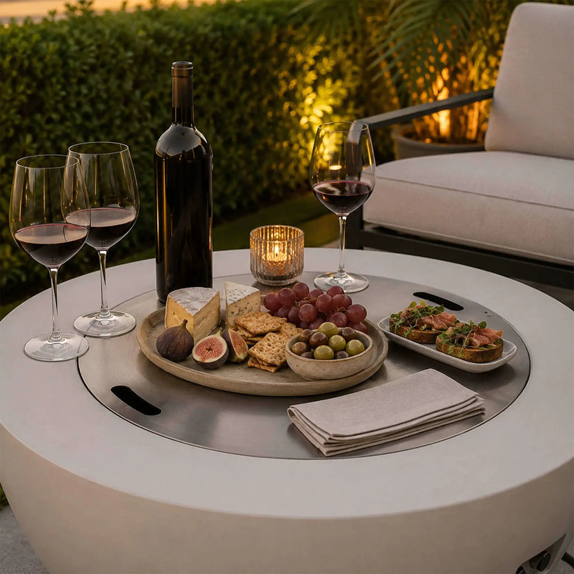 Lifestyle image of the Flame Havens Refuge Series 36-inch Round Natural Gas White GFRC Concrete Fire Pit with the brushed stainless steel burner cover installed and used as an outdoor entertaining table. A wine bottle, glasses, artisan cheeses, fruit, appetizers, and candle create an elegant gathering space for luxury outdoor living.