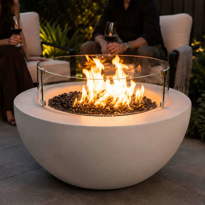 Lifestyle image of the Flame Havens Refuge Series 36-inch Round Natural Gas White GFRC Concrete Fire Pit featuring the optional tempered glass wind guard. Friends relax around the glowing fire on a luxury outdoor patio, highlighting how the glass wind guard protects the flames while creating a warm, inviting gathering space for evening entertaining.