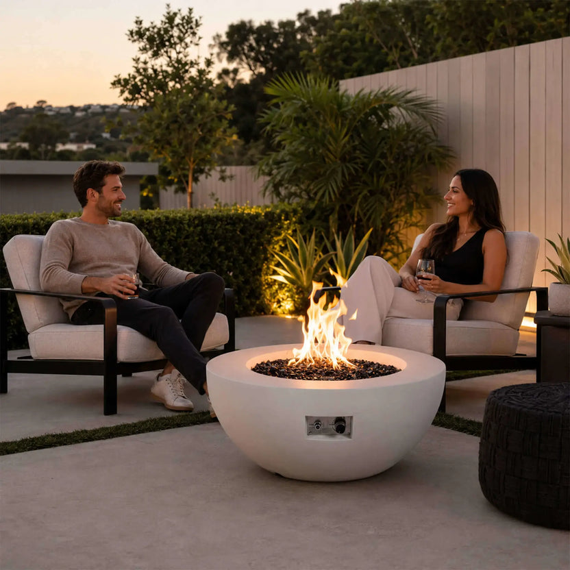 The Flame Havens Refuge Series 36-inch Round Natural Gas White GFRC Concrete Fire Pit serves as the focal point of a sophisticated backyard entertaining space. A couple relaxes in modern outdoor lounge chairs while enjoying the warm flames, demonstrating how the premium white concrete fire pit enhances luxury patios, outdoor living rooms, and evening gatherings.