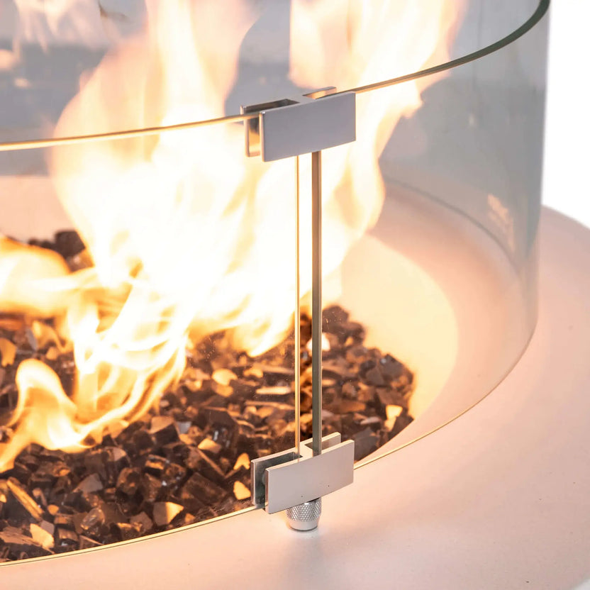 Close-up detail of the Flame Havens Refuge Series 36-inch Round Natural Gas White GFRC Concrete Fire Pit highlighting the optional tempered glass wind guard hardware. The image showcases the precision-machined stainless steel mounting brackets, secure glass attachment system, premium black reflective tempered fire glass, and vibrant natural gas flames for a refined, high-end outdoor fire feature.