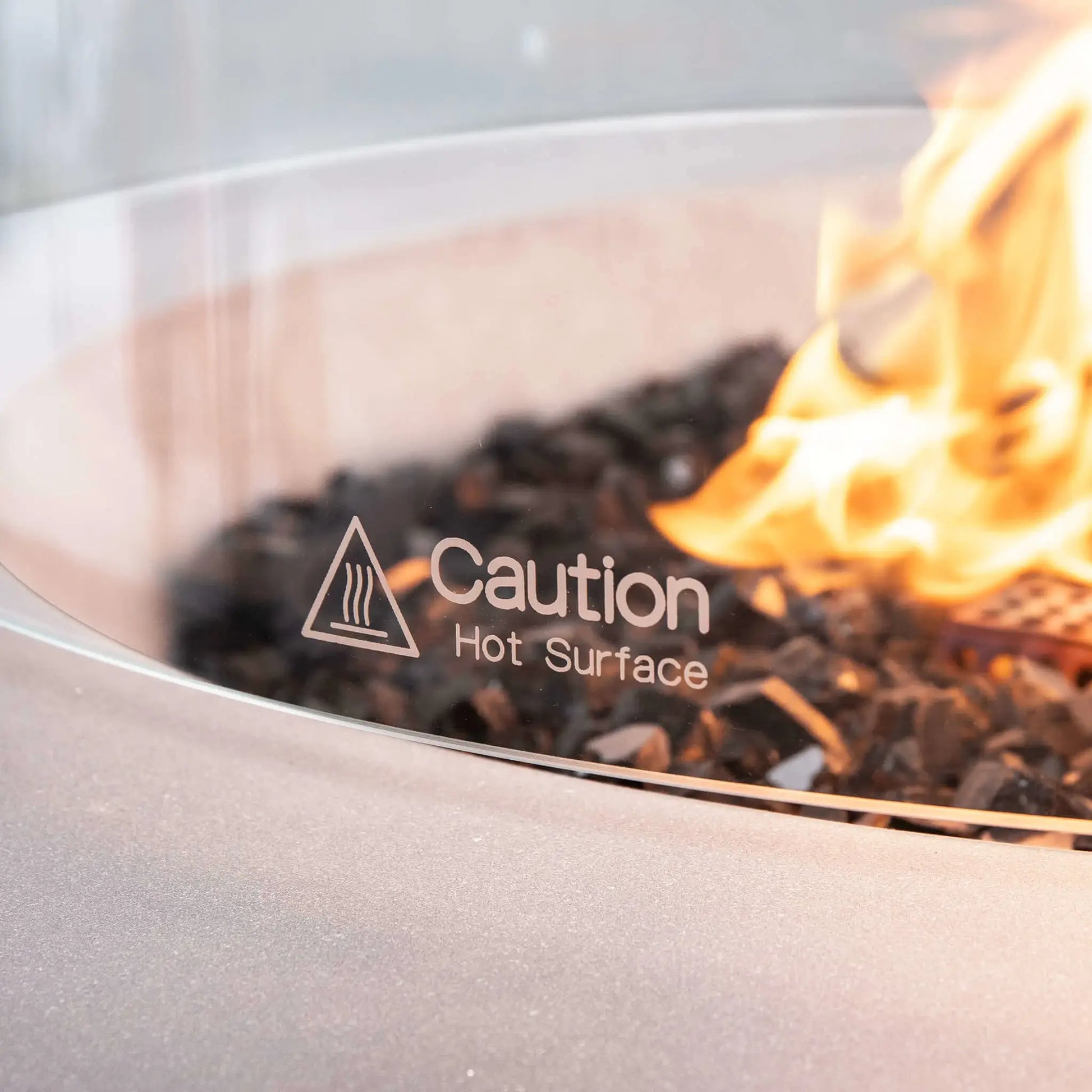 Macro close-up of the Flame Havens Refuge Series 36-inch Round Natural Gas White GFRC Concrete Fire Pit tempered glass wind guard featuring the etched "Caution Hot Surface" safety marking. Warm natural gas flames illuminate the premium black reflective tempered fire glass while the crystal-clear tempered glass demonstrates the wind guard's safety, durability, and premium finish for luxury outdoor living spaces.