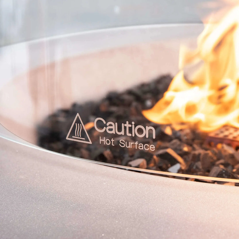 Macro close-up of the Flame Havens Refuge Series 36-inch Round Natural Gas White GFRC Concrete Fire Pit tempered glass wind guard featuring the etched "Caution Hot Surface" safety marking. Warm natural gas flames illuminate the premium black reflective tempered fire glass while the crystal-clear tempered glass demonstrates the wind guard's safety, durability, and premium finish for luxury outdoor living spaces.