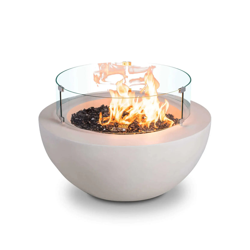 Front studio image of the Flame Havens Refuge Series 36-inch Round Natural Gas White GFRC Concrete Fire Pit operating with the optional tempered glass wind guard installed. Bright flames rise above premium black reflective tempered fire glass while the clear cylindrical wind guard with stainless steel mounting brackets enhances safety, flame stability, and the modern architectural appearance of this luxury outdoor fire pit.