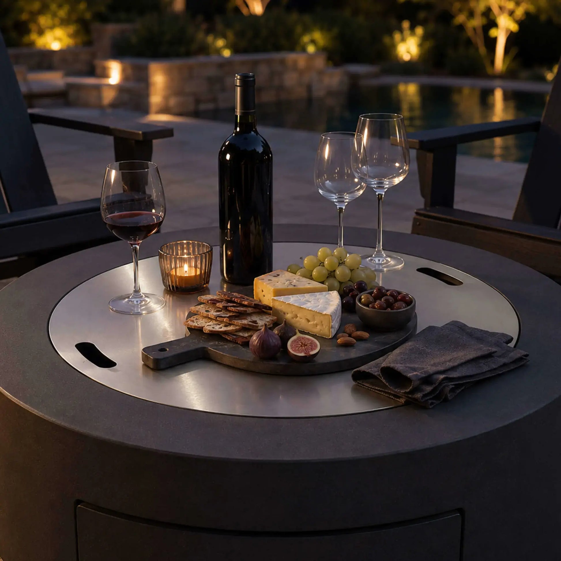 Lifestyle image of the Flame Havens Refuge Series 36-inch Round Propane Gas GFRC Concrete Fire Pit with the optional Stainless Steel Burner Cover installed and used as an elegant outdoor tabletop. Displayed with wine, cheese, fruit, crackers, and candles beside a luxury pool, the Graphite Black GFRC fire pit seamlessly combines outdoor entertaining with premium functionality.