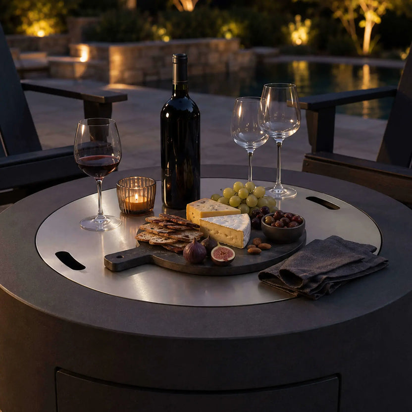 Lifestyle image of the Flame Havens Refuge Series 36-inch Round Propane Gas GFRC Concrete Fire Pit with the optional Stainless Steel Burner Cover installed and used as an elegant outdoor tabletop. Displayed with wine, cheese, fruit, crackers, and candles beside a luxury pool, the Graphite Black GFRC fire pit seamlessly combines outdoor entertaining with premium functionality.