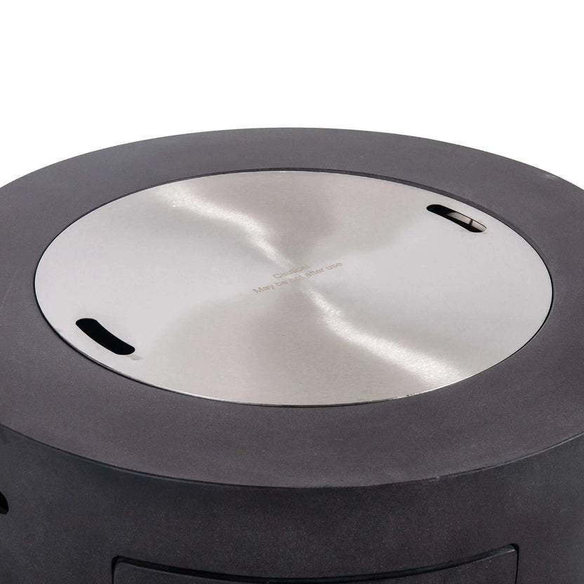 Flame Havens Refuge Series 36-inch Round Propane Gas GFRC Concrete Fire Pit in Graphite Black featuring the optional Stainless Steel Burner Cover installed flush within the burner opening. The brushed stainless steel cover transforms the fire pit into a functional tabletop while protecting the burner from weather, debris, and moisture when the fire feature is not in use.