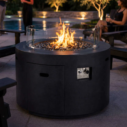 Lifestyle image of the Flame Havens Refuge Series 36-inch Round Propane Gas GFRC Concrete Fire Pit with premium Black Reflective Fire Glass and the optional tempered glass wind guard installed. Set beside a luxury backyard pool and surrounded by Adirondack chairs, the Graphite Black GFRC fire pit creates a warm, inviting focal point for outdoor entertaining while protecting the flame from wind.