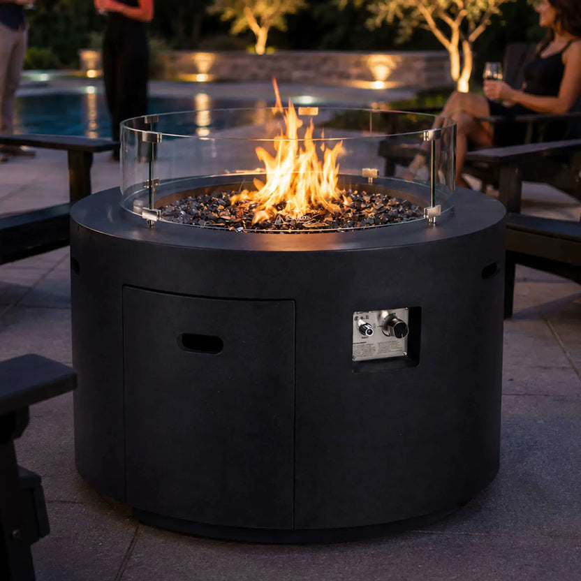 Lifestyle image of the Flame Havens Refuge Series 36-inch Round Propane Gas GFRC Concrete Fire Pit with premium Black Reflective Fire Glass and the optional tempered glass wind guard installed. Set beside a luxury backyard pool and surrounded by Adirondack chairs, the Graphite Black GFRC fire pit creates a warm, inviting focal point for outdoor entertaining while protecting the flame from wind.