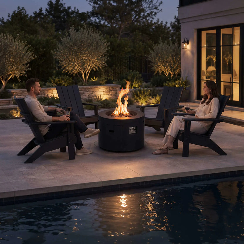 Lifestyle image of the Flame Havens Refuge Series 36-inch Round Propane Gas GFRC Concrete Fire Pit in Graphite Black surrounded by Adirondack chairs beside a luxury backyard pool. A couple relaxes around the fire while enjoying drinks, showcasing the propane fire pit's natural flames, Black Reflective Fire Glass, recessed controls, and inviting atmosphere for outdoor entertaining.