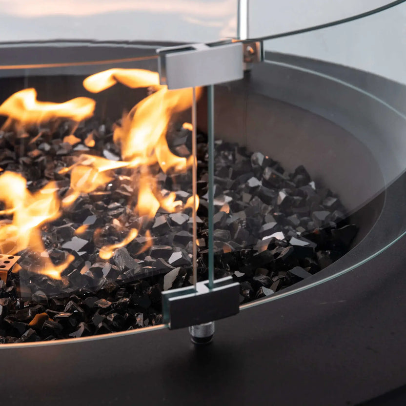 Detailed close-up of the Flame Havens Refuge Series 36-inch Round Propane Gas GFRC Concrete Fire Pit optional tempered glass wind guard. The image showcases the precision stainless steel mounting brackets, clear tempered glass panels, premium Black Reflective Fire Glass, and natural propane flames, highlighting the craftsmanship and secure installation of the wind guard accessory.
