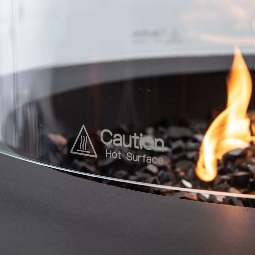 Close-up of the Flame Havens Refuge Series 36-inch Round Propane Gas GFRC Concrete Fire Pit tempered glass wind guard featuring the etched "Caution Hot Surface" safety marking. The image highlights the crystal-clear tempered glass, premium Black Reflective Fire Glass, and natural propane flame while showcasing the safety features and high-quality construction of the optional wind guard accessory.