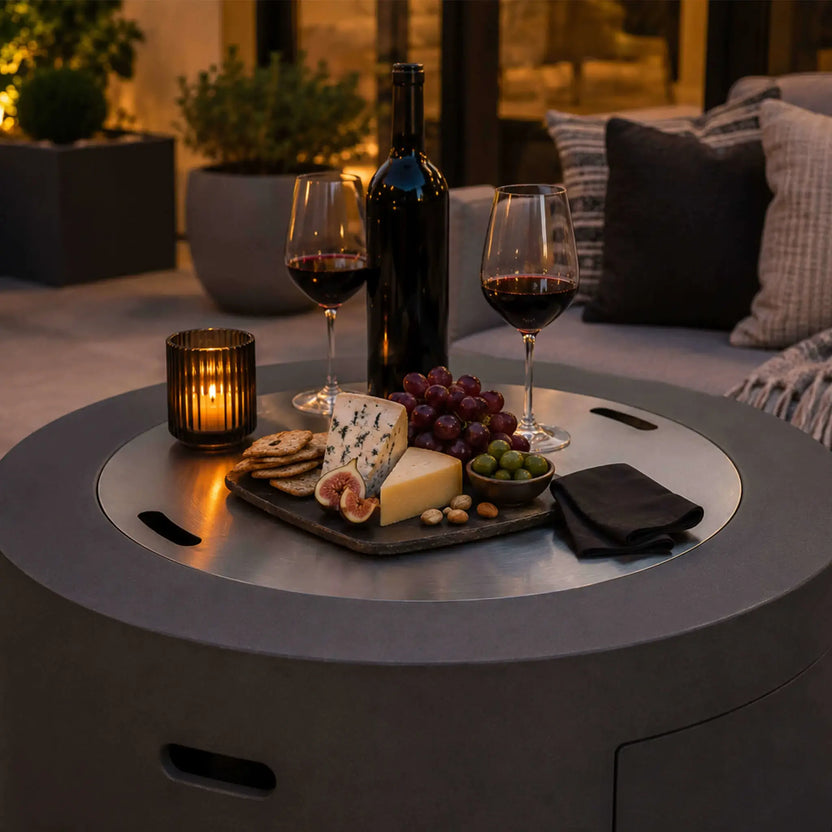 Lifestyle image of the Flame Havens Refuge Series 36-inch Round Propane Gas GFRC Concrete Fire Pit in Natural Gray with the optional Stainless Steel Burner Cover installed, transforming the fire pit into an elegant outdoor table. A charcuterie board, wine glasses, candle, and bottle of wine create a sophisticated entertaining space on a luxury patio, highlighting the versatility of this propane gas fire pit.