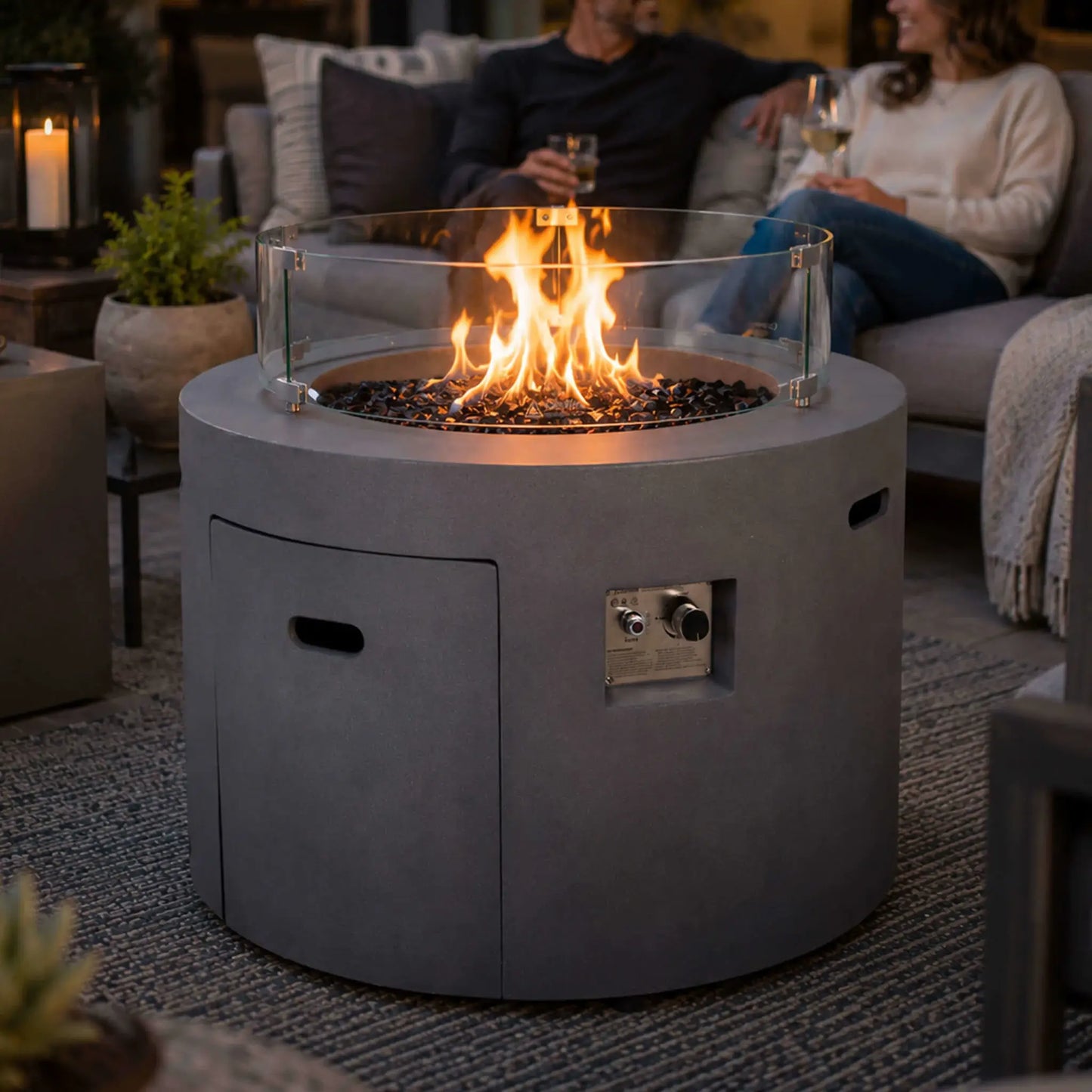 Lifestyle image of the Flame Havens Refuge Series 36-inch Round Propane Gas GFRC Concrete Fire Pit in Natural Gray with premium Black Reflective Fire Glass and the optional tempered glass wind guard. Set within a luxury outdoor seating area, the fire pit provides warmth and ambiance while family and friends relax around the modern GFRC concrete centerpiece.
