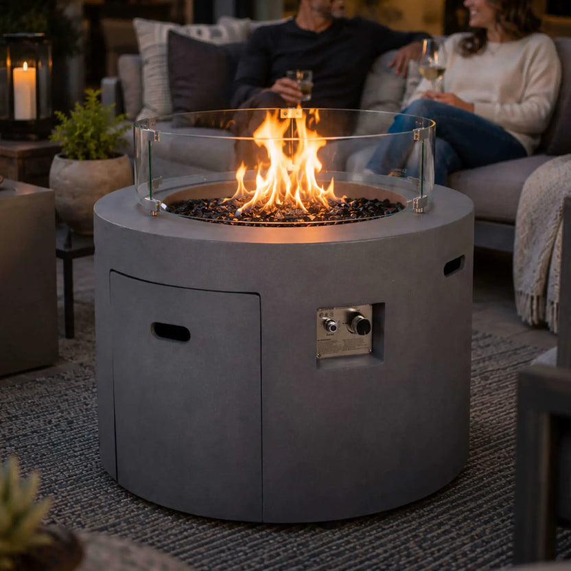 Lifestyle image of the Flame Havens Refuge Series 36-inch Round Propane Gas GFRC Concrete Fire Pit in Natural Gray with premium Black Reflective Fire Glass and the optional tempered glass wind guard. Set within a luxury outdoor seating area, the fire pit provides warmth and ambiance while family and friends relax around the modern GFRC concrete centerpiece.