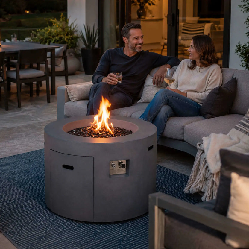 Lifestyle image of the Flame Havens Refuge Series 36-inch Round Propane Gas GFRC Concrete Fire Pit in Natural Gray burning with vibrant propane flames above Black Reflective Fire Glass. Positioned in a luxury backyard patio, the fire pit creates a warm gathering space as a couple enjoys drinks on a modern outdoor sofa, demonstrating the elegant design and inviting ambiance of the Refuge Series.