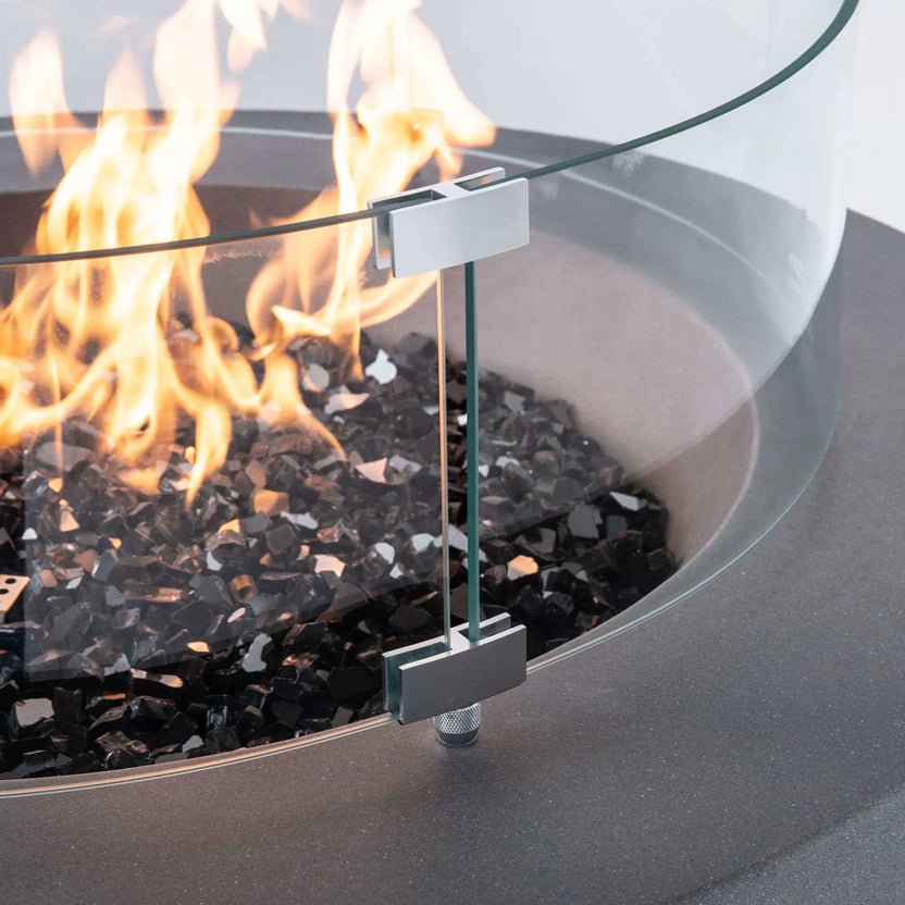 Close-up of the optional tempered glass wind guard installed on the Flame Havens Refuge Series 36-inch Round Propane Gas GFRC Concrete Fire Pit. The image highlights the precision stainless steel mounting brackets, crystal-clear tempered glass panels, vibrant propane flames, and premium Black Reflective Fire Glass, showcasing the wind guard's secure construction and elegant appearance.