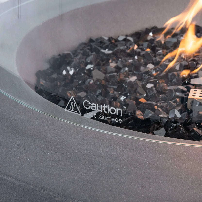 Detailed close-up of the tempered glass wind guard on the Flame Havens Refuge Series 36-inch Round Propane Gas GFRC Concrete Fire Pit featuring premium Black Reflective Fire Glass and active propane flames. The image showcases the etched "Caution Hot Surface" safety marking on the tempered glass, emphasizing the premium finish, safety features, and durable outdoor design of the optional wind guard accessory.