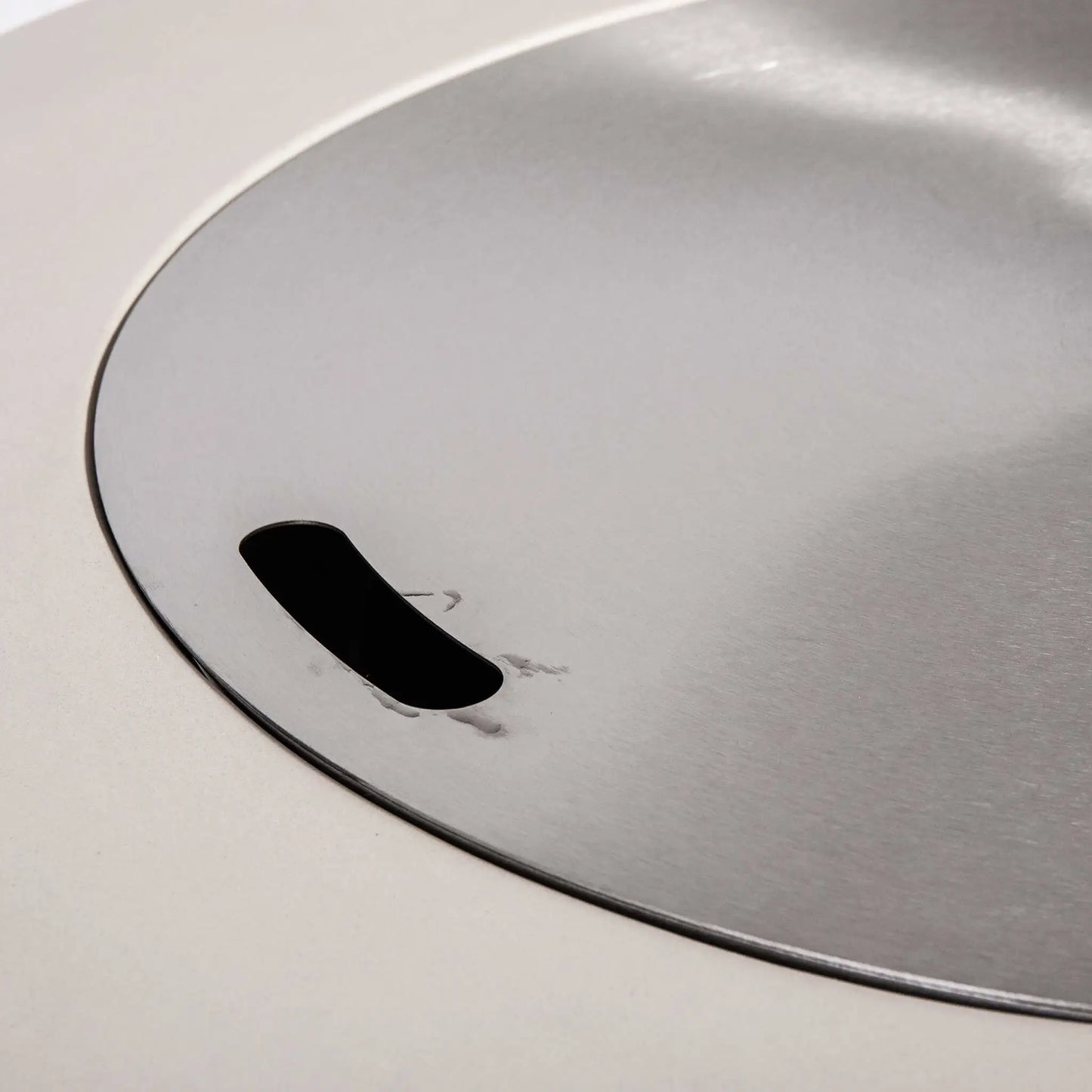 Close-up of the optional Stainless Steel Burner Cover for the Flame Havens Refuge Series 36-inch Round Propane Gas GFRC Concrete Fire Pit in Ivory White. The brushed stainless steel cover sits flush within the burner opening and features an integrated finger slot for easy removal, transforming the fire pit into a functional tabletop when the burner is not in use.