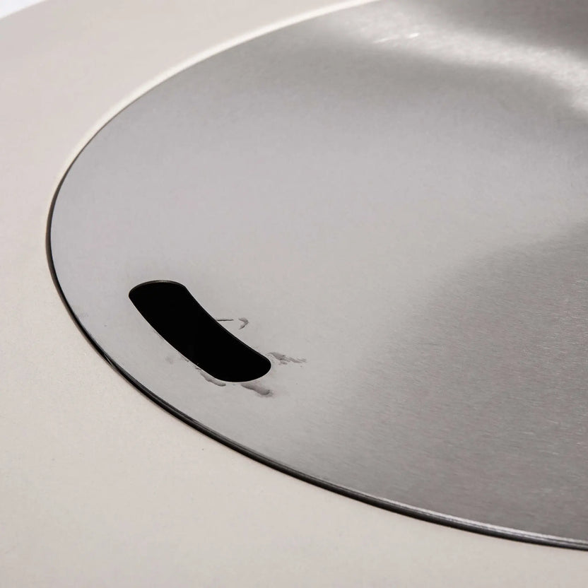 Close-up of the optional Stainless Steel Burner Cover for the Flame Havens Refuge Series 36-inch Round Propane Gas GFRC Concrete Fire Pit in Ivory White. The brushed stainless steel cover sits flush within the burner opening and features an integrated finger slot for easy removal, transforming the fire pit into a functional tabletop when the burner is not in use.