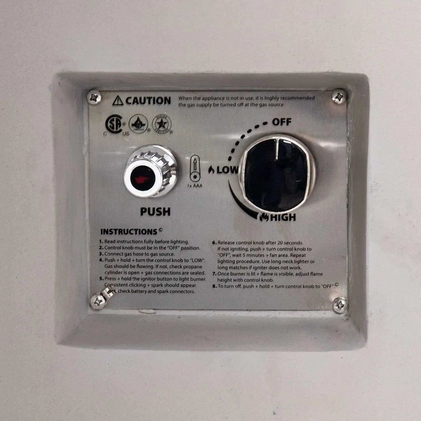Close-up of the CSA-certified ignition control panel on the Flame Havens Refuge Series 36-inch Round Propane Gas GFRC Concrete Fire Pit in Ivory White. The recessed stainless steel panel features the push-button igniter, flame adjustment control knob, OFF-LOW-HIGH settings, lighting instructions, and safety certifications for reliable propane gas operation.