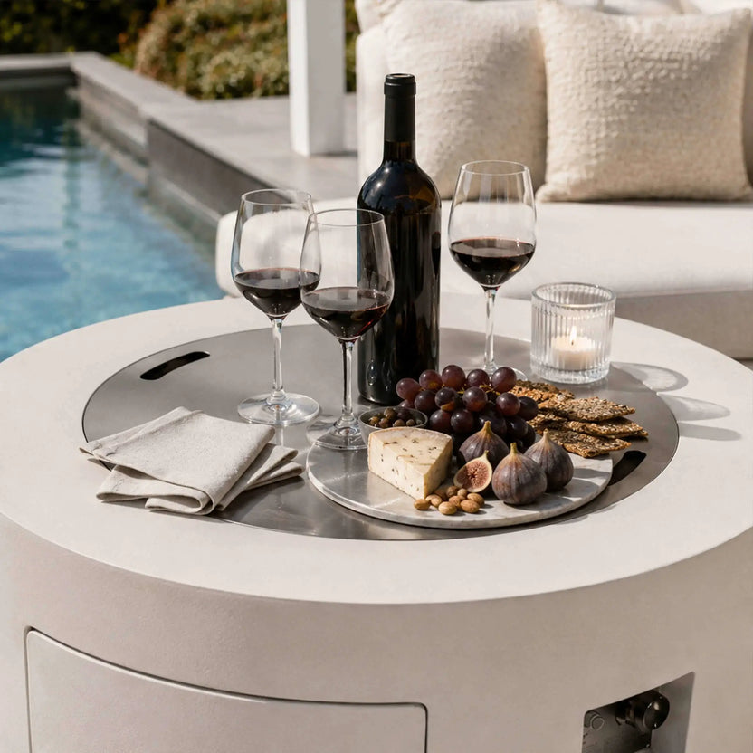 Lifestyle image of the Flame Havens Refuge Series 36-inch Round Propane Gas GFRC Concrete Fire Pit in Ivory White with the optional Stainless Steel Burner Cover installed and used as a tabletop. The cover supports a wine bottle, glasses, cheese board, fruit, crackers, and candle beside a luxury pool, demonstrating how the fire pit easily transitions into an elegant outdoor entertaining table when the burner is covered.