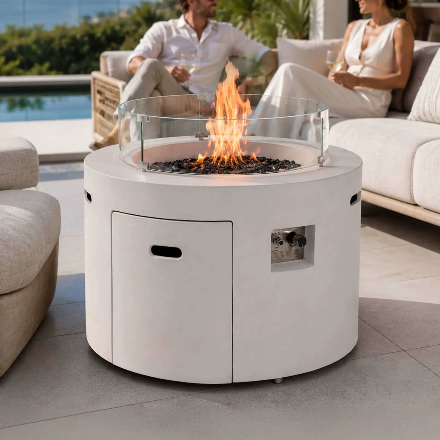 Lifestyle image of the Flame Havens Refuge Series 36-inch Round Propane Gas GFRC Concrete Fire Pit in Ivory White featuring premium Black Reflective Fire Glass and an optional tempered glass wind guard. Positioned on a luxury outdoor patio beside comfortable seating, the fire pit creates a warm focal point while complementing elegant modern outdoor living spaces.